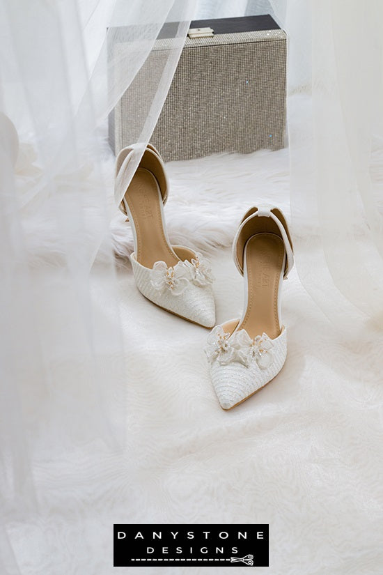 White bridal shoes with chiffon butterfly wings and 9cm heels, displayed on a white surface with a bridal bag and flowers in the background.