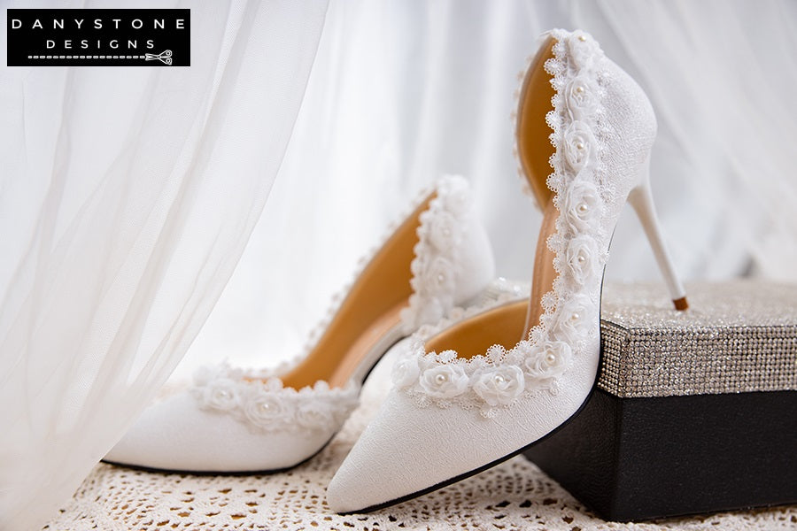 Pair of white wedding shoes with rose lace trim and 9cm heels, photographed outdoors with a natural background.