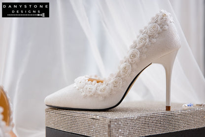 Pair of white wedding shoes with rose lace trim and 9cm heels, displayed on a glittery box with a lace fabric background.