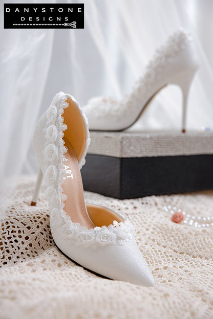 Elegant white wedding shoes with rose lace trim, displayed on a glittery box against a sheer fabric backdrop.