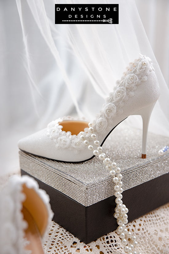 Elegant white wedding shoes with rose lace trim, displayed on a glittery box against a sheer fabric backdrop.