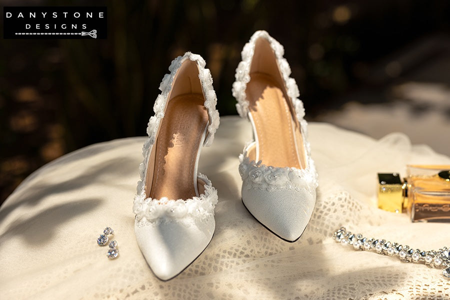 Pair of white wedding shoes with rose lace trim, displayed on a lace-covered table with jewelry and a perfume bottle.