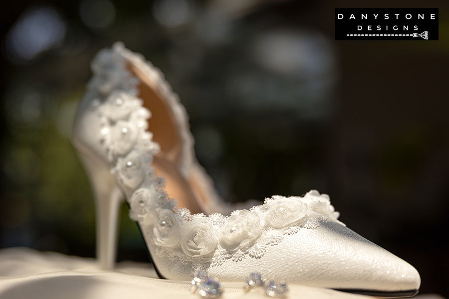 Pair of white wedding shoes with rose lace trim, displayed on a lace-covered table with jewelry and a perfume bottle.