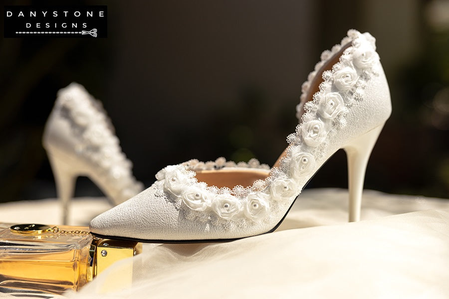 Pair of white wedding shoes with rose lace trim, displayed on a lace-covered table with jewelry and a perfume bottle.