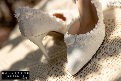 Close-up of a white wedding shoe with rose lace trim and 9cm heel, showcased with sparkling jewelry in the foreground.