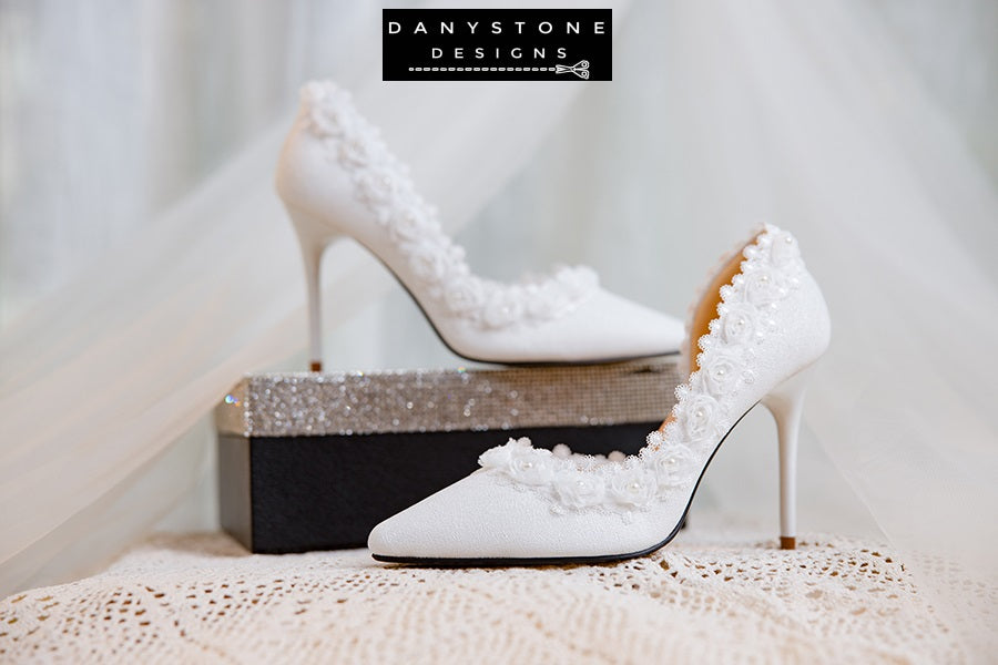 Side view of a white wedding shoe with metallic scratched leather and rose lace trim, displayed with a perfume bottle.