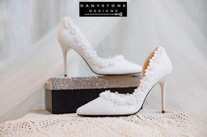 Side view of a white wedding shoe with metallic scratched leather and rose lace trim, displayed with a perfume bottle.