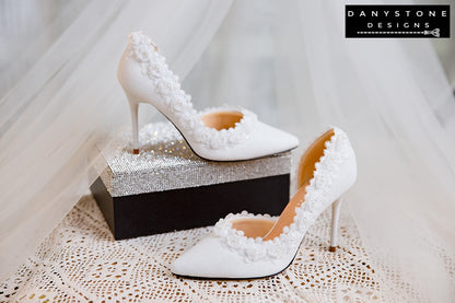 Side view of a white wedding shoe with metallic scratched leather and rose lace trim, displayed with a perfume bottle.