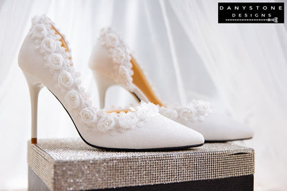 Side view of a white wedding shoe with metallic scratched leather and rose lace trim, displayed with a perfume bottle.