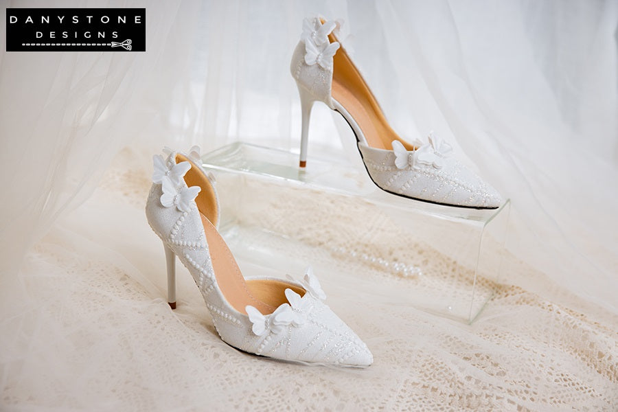 Pair of white lace wedding shoes with butterfly decor, displayed against a glittery box with a sheer fabric backdrop.