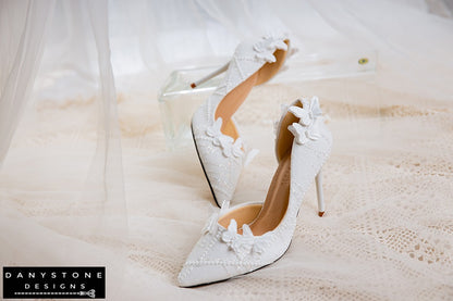 White lace wedding shoes with butterfly decor, shown on a lace-covered surface with a glass stand.