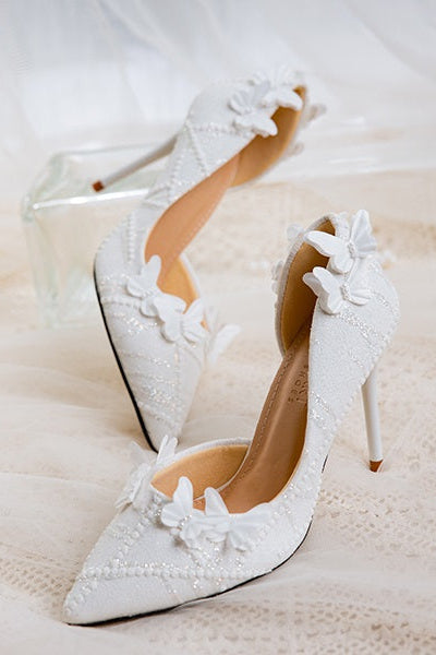 White lace wedding shoes with butterfly decor, shown on a lace-covered surface with a glass stand.