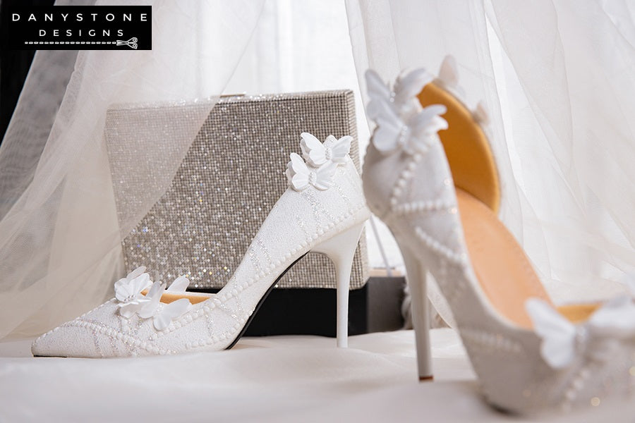 Pair of white lace wedding shoes with butterfly decor, displayed on a rustic table with a floral arrangement in the background.