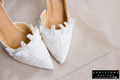 Pair of white lace wedding shoes with butterfly decor, displayed on a rustic table with a floral arrangement in the background.