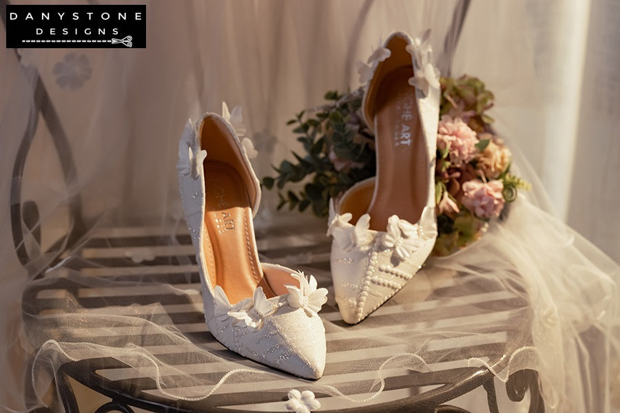 Pair of white lace wedding shoes with butterfly decor, elegantly arranged on a table with a floral bouquet in the background.