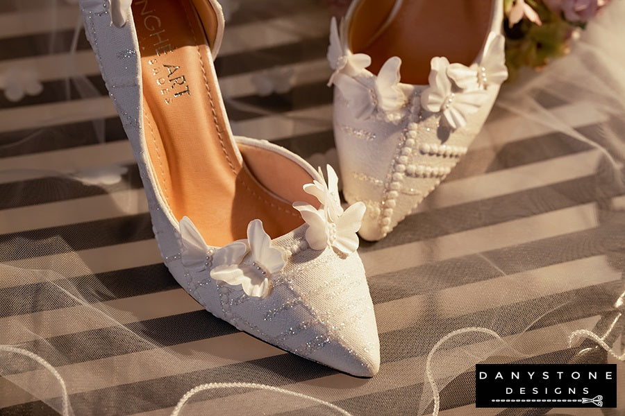 Pair of white lace wedding shoes with butterfly decor, elegantly arranged on a table with a floral bouquet in the background.