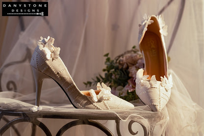 White lace wedding shoes with butterfly decor, shown on a lace-covered surface with a glass stand.