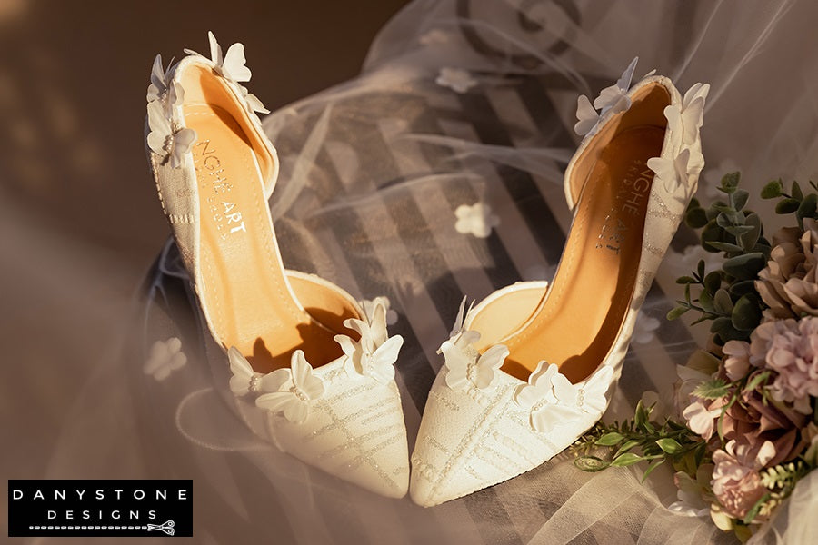 Side view of white lace wedding shoes with butterfly decor and 9cm heels, displayed with a glittery box and a sheer fabric backdrop.