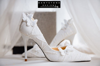 Pair of white lace wedding shoes with butterfly decor, displayed against a glittery box with a sheer fabric backdrop.