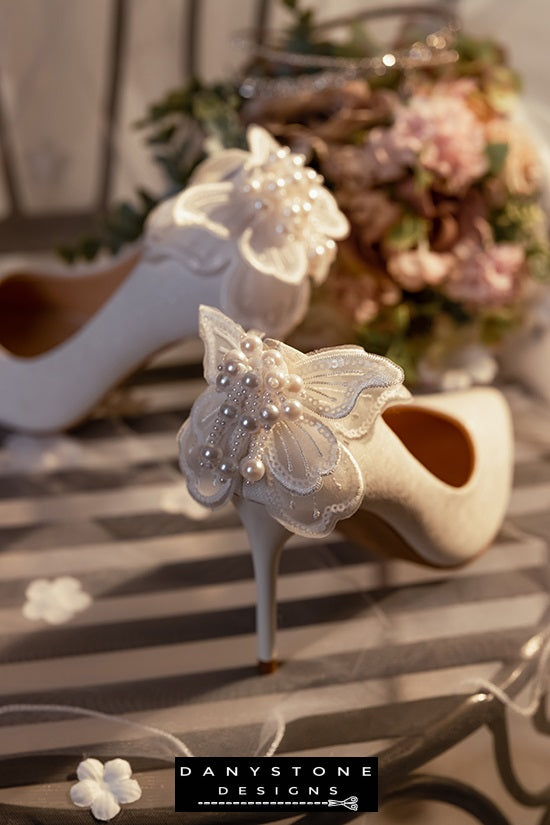 White wedding shoes with butterfly wings, displayed on a lace-covered chair with a floral arrangement in the background.