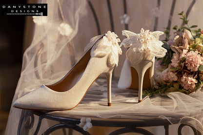 White wedding shoes with butterfly wings, displayed on a lace-covered chair with a floral arrangement in the background.