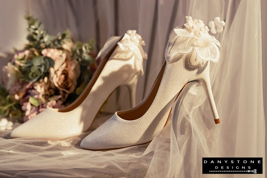 Pair of white wedding shoes with butterfly wings, shown in a romantic setting with flowers and tulle.