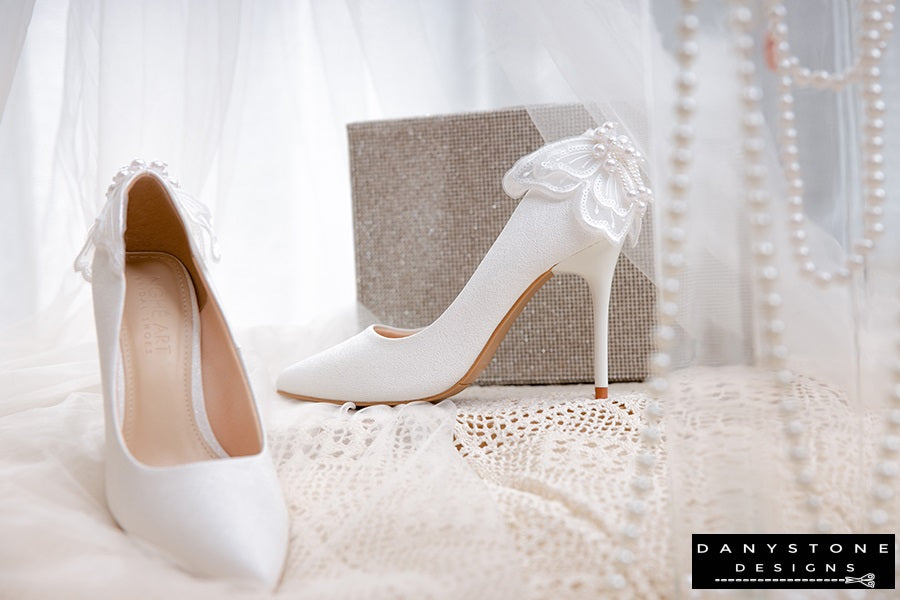 Pair of white wedding shoes with butterfly wings, featured with a pearl necklace and a glittery box.