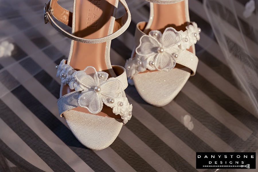 Elegant White Floral Wedding High Heels on a wooden surface with rolled-up white fabric in the background.