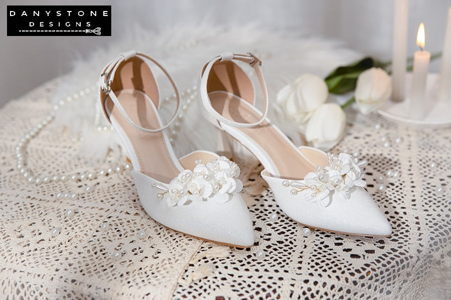 Pair of Elegant White Floral Wedding Shoes with floral patterns and ankle straps, showcased on a lace tablecloth with white roses.