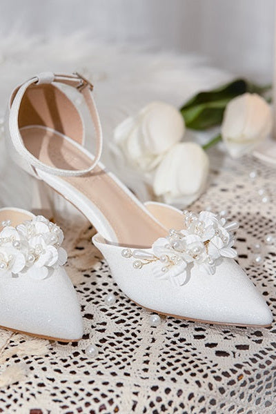 Pair of Elegant White Floral Wedding Shoes with floral patterns and ankle straps, showcased on a lace tablecloth with white roses.