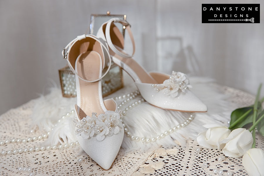 Elegant White Floral Wedding Shoes with an ankle strap on a lace tablecloth, featuring lightly shimmering metallic leather and floral patterns.