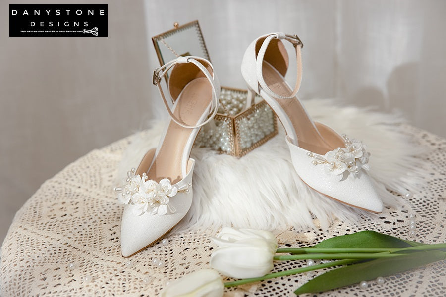 Elegant White Floral Wedding Shoes with floral patterns and ankle straps, displayed with pearls and flowers in the background.