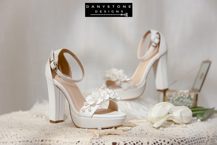 Pair of Elegant White Matte Leather Wedding Sandals with floral embellishments on a lace tablecloth.