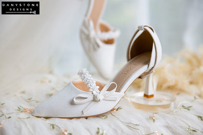 Top view of white bridal shoes with floral straps and bows, 7cm heels, arranged on a floral fabric background.