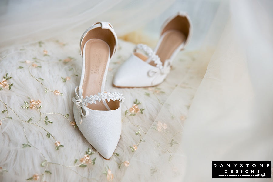 Side view of white bridal shoes with floral straps and bows, 7cm heels, displayed on a floral fabric background.