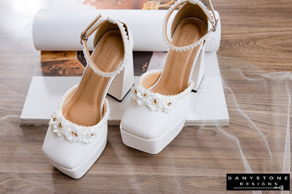 White wedding shoes with double square toe, 13cm heel, floral decorations, and pearl trim, arranged on a wooden surface.