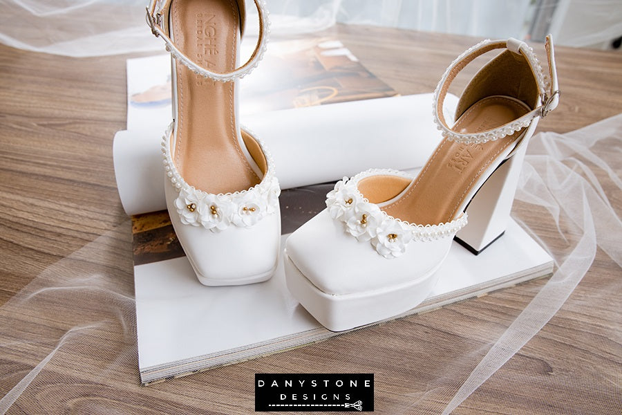 Stylish white bridal shoes with double square toe, 13cm heel, and floral trim, displayed on a book.