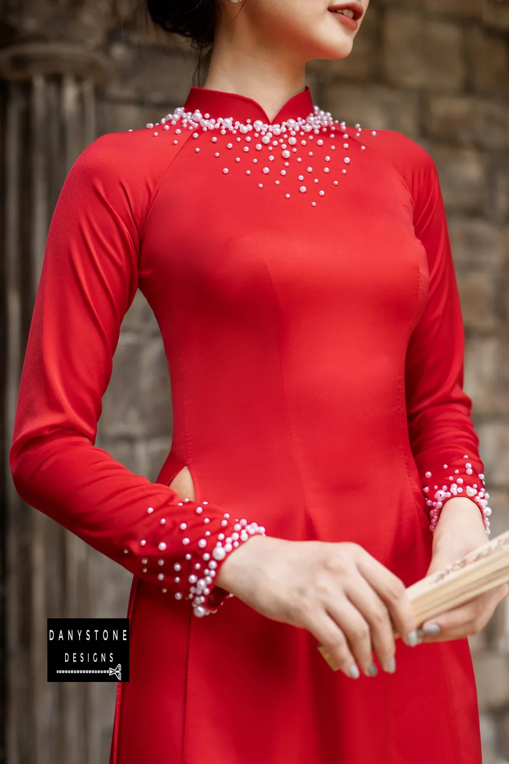 Elegant woman wearing a red silk Áo Dài with pearl details, holding a fan.