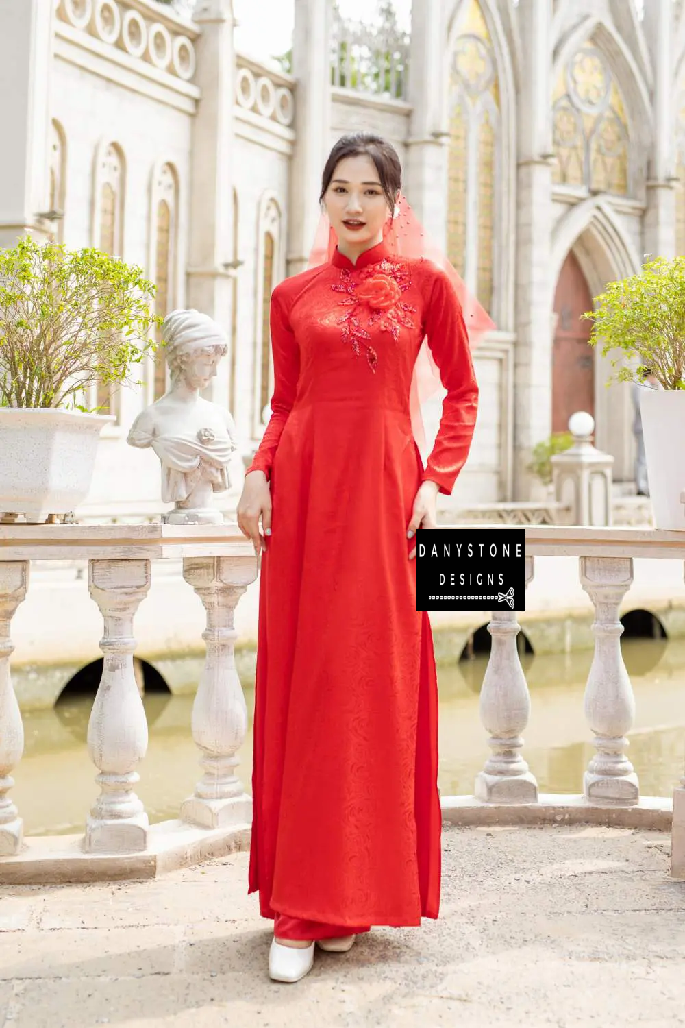 Full-length view of a bride in a red brocade Áo Dài with 4 panels and a large floral design.