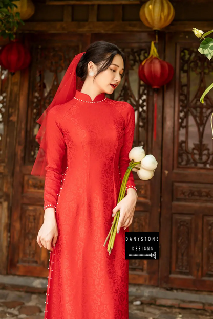 Woman wearing a red brocade Áo Dài with 4 panels and floral background, smiling while holding white flowers.