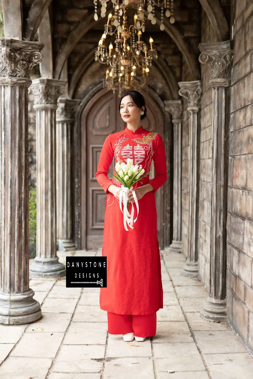 Elegant woman in a royal red brocade bridal Áo Dài with phoenix embroidery, holding a bouquet.