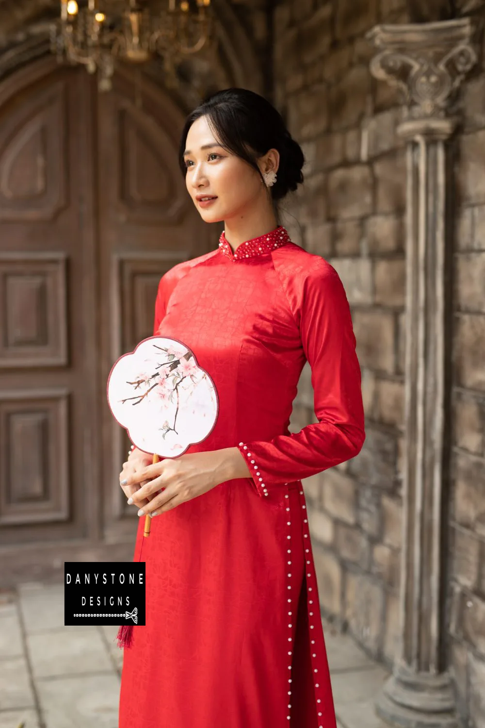 Full-length view of a bride in a red brocade Áo Dài with pearls on the sleeves and hem.