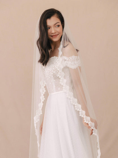 Woman wearing a white lace wedding dress with a matching veil against a beige background