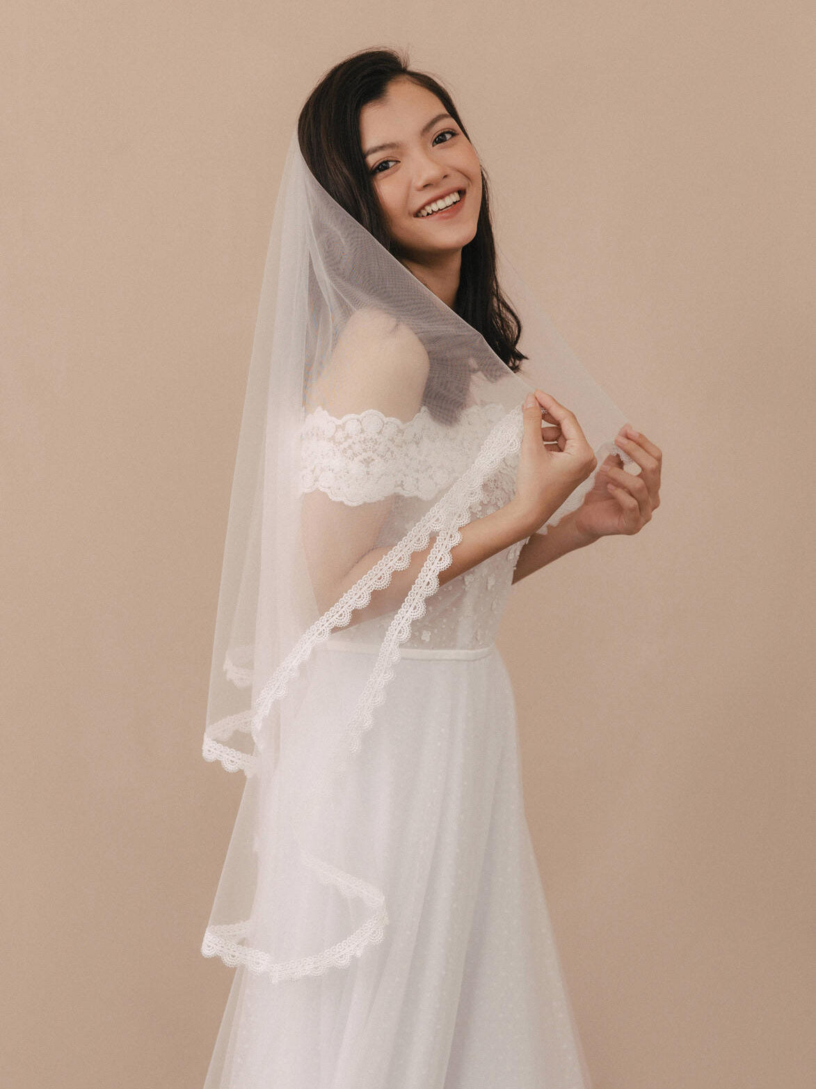 Woman wearing a white lace wedding dress with a matching veil against a beige background