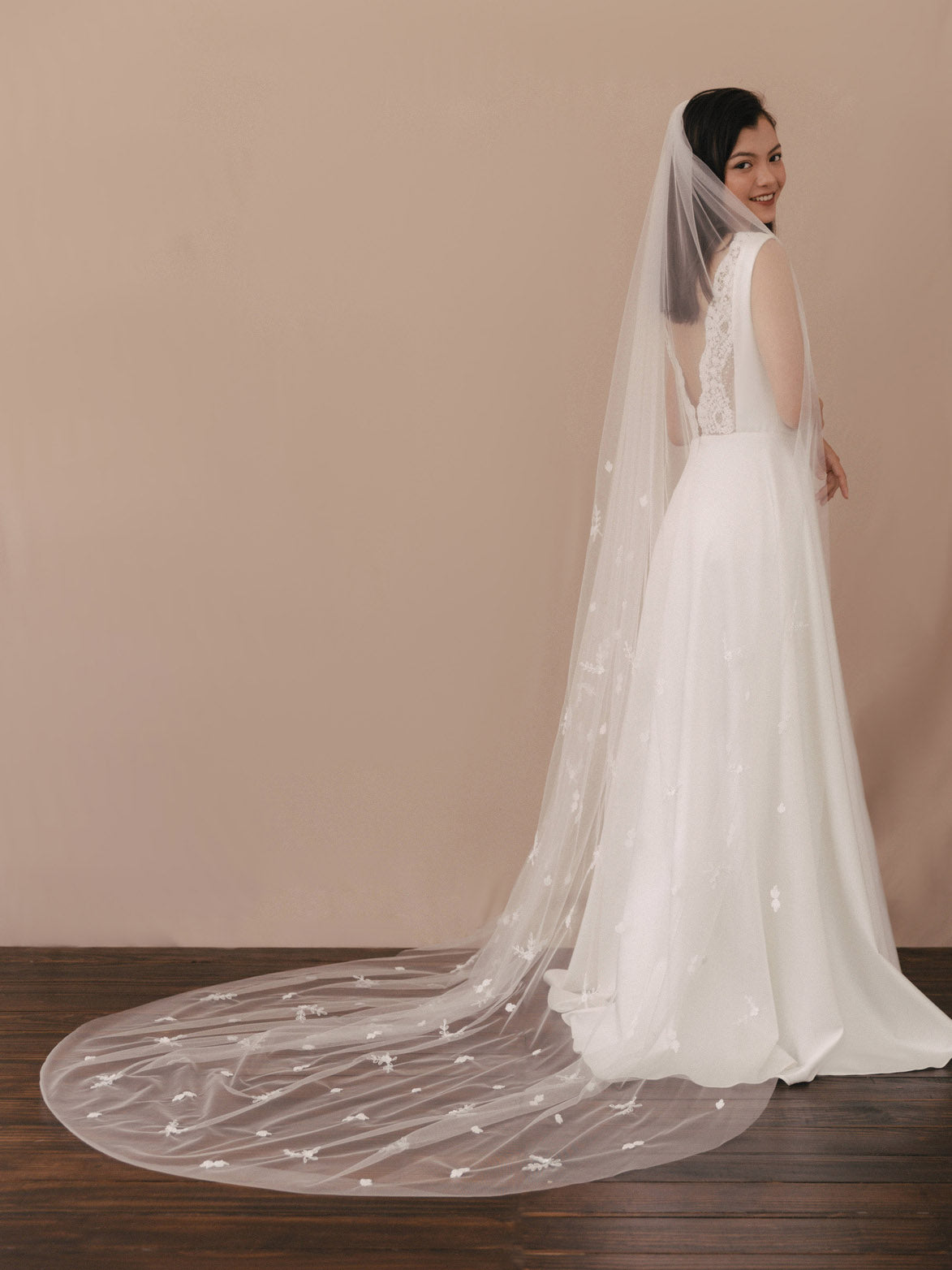 Woman wearing a white wedding dress with a long veil against a beige wall.