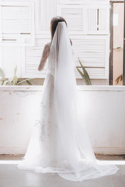 Person wearing a long white wedding veil in front of a white wall with plants.