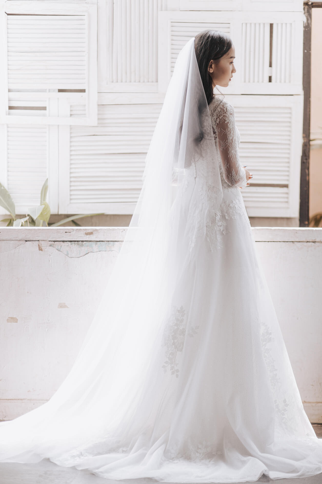 Woman in a white wedding dress with a long veil standing in front of a window.