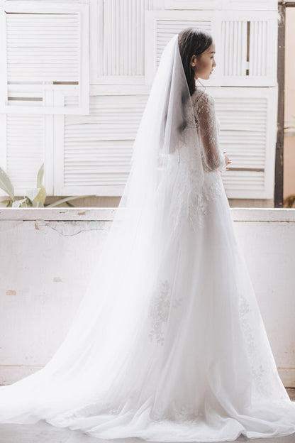 Woman in a white wedding dress with a long veil standing in front of a window.