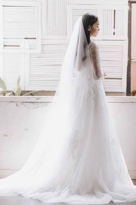 Woman in a white wedding dress with a long veil standing in front of a window.