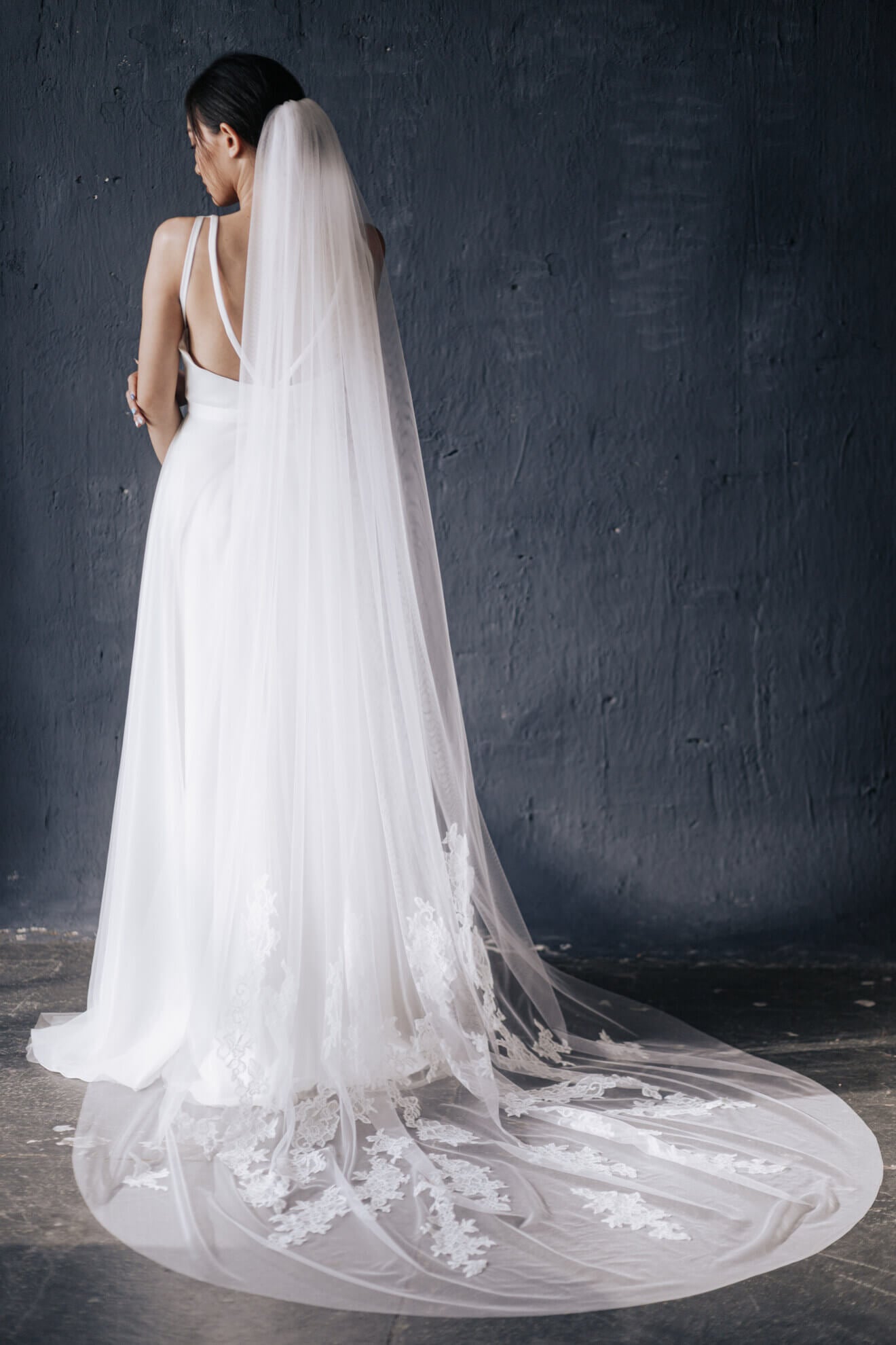 Bride wearing a long, elegant white wedding dress with a matching veil against a dark background.